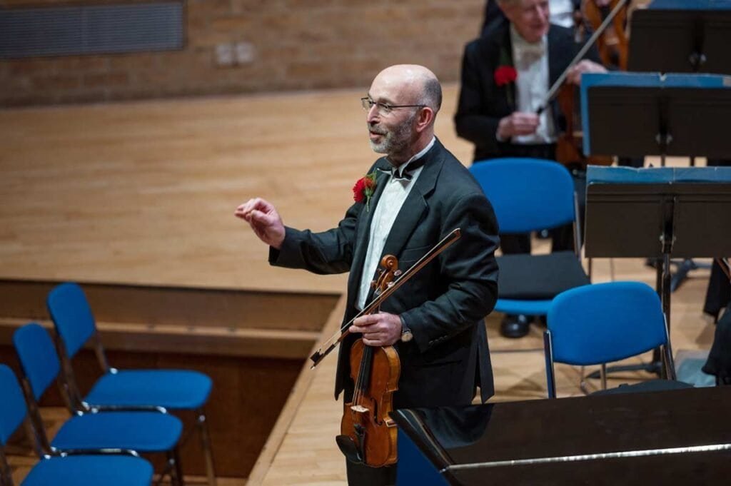 Acoustic violinist conducting Sampson Orchestra during a classical performance in a concert hall.